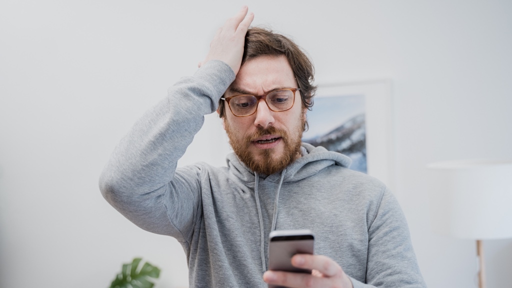 En man i grå hoodie ser oroad ut när han tittar på sin telefon och håller ena handen mot huvudet i bekymmer.