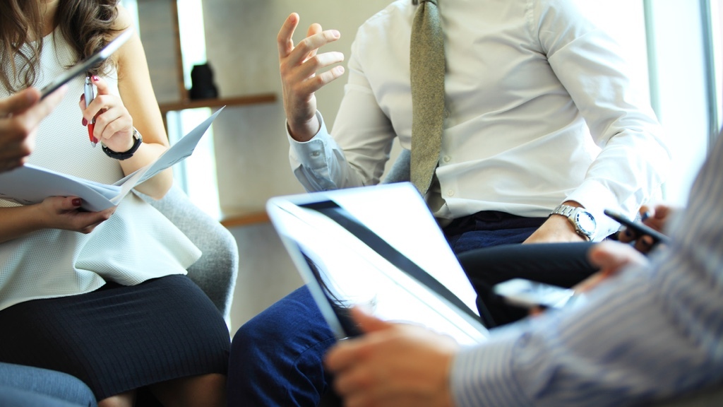 En grupp yrkespersoner har ett affärsmöte, där de håller i dokument, surfplattor och mobiltelefoner medan de engagerat diskuterar något.