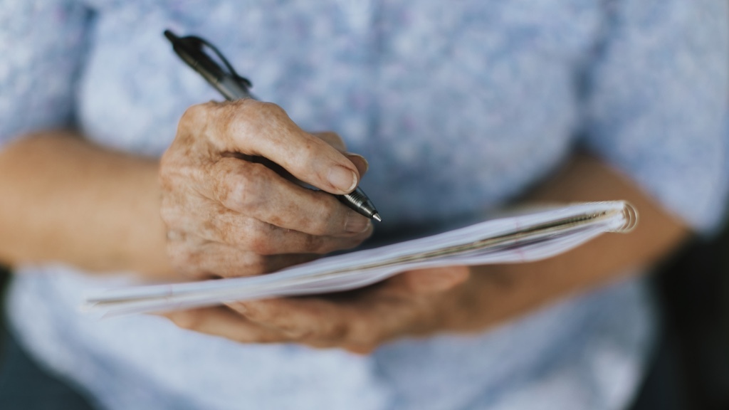 En äldre person skriver i ett anteckningsblock med en penna, hållandes det nära bröstet.