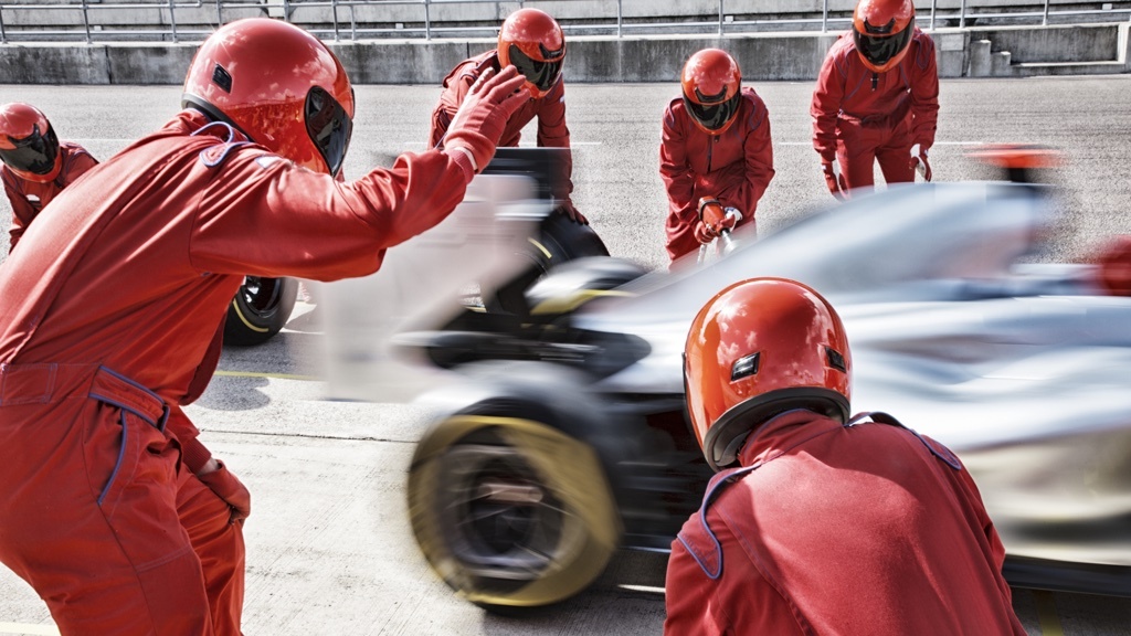 A Formula 1 pit crew in red suits prepares to service a race car speeding into the pit lane.
