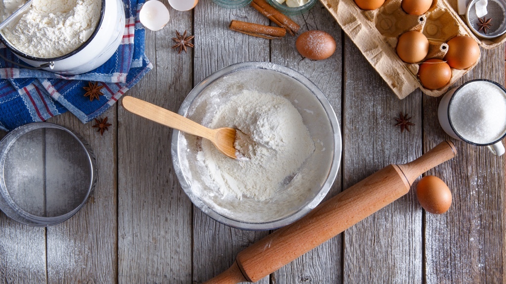 Ingredienser och redskap för bakning, inklusive mjöl, ägg, socker, kanelstänger och en kavel, är prydligt upplagda på ett rustikt träbord.