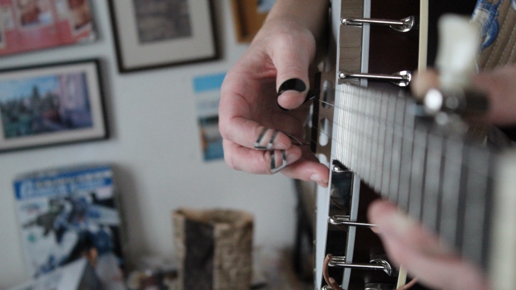En person spelar banjo med metallplektrum på fingrarna, och handen är fångad i närbild mot en bakgrund av inramade bilder och affischer.