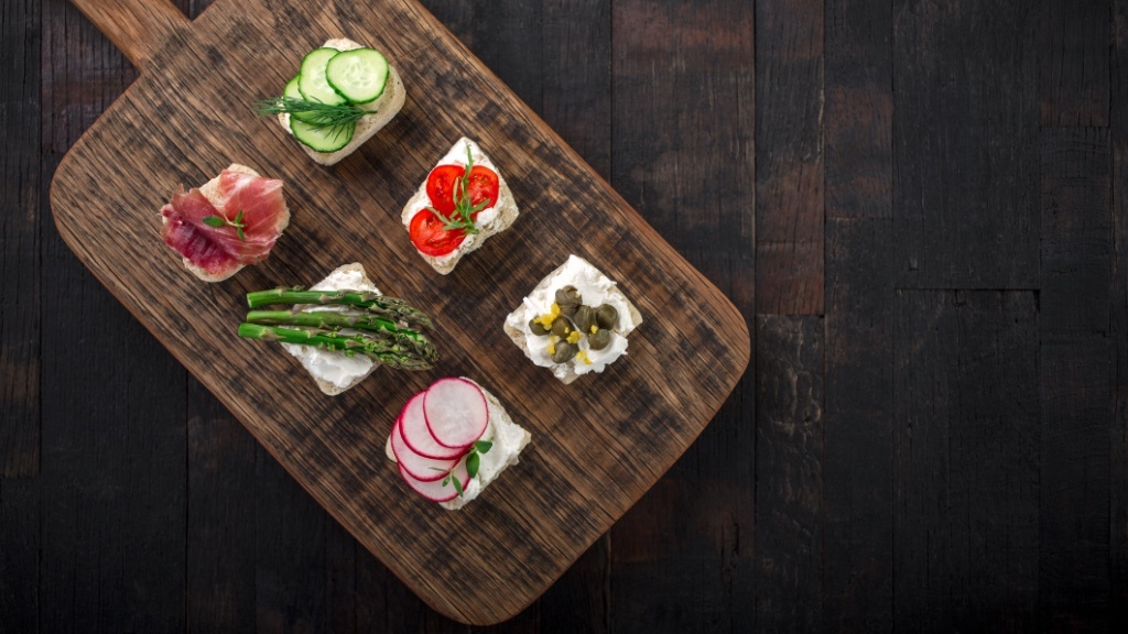 Six small, decorative appetizers with various toppings like cucumber, tomato, asparagus, radish, prosciutto, and capers are arranged on a wooden serving board.