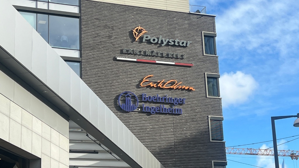 En husfasad visar företagsloggor för bland annat Polystar, Erik Olsson och Boehringer Ingelheim, under en blå himmel där en byggkran syns i bakgrunden.