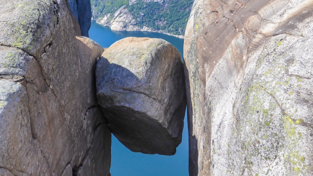 Ett stort stenblock sitter fastkilat mellan två höga klippor ovanför en djupblå fjord.