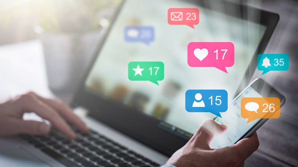 A person uses a smartphone and laptop simultaneously, surrounded by floating social media notification icons.