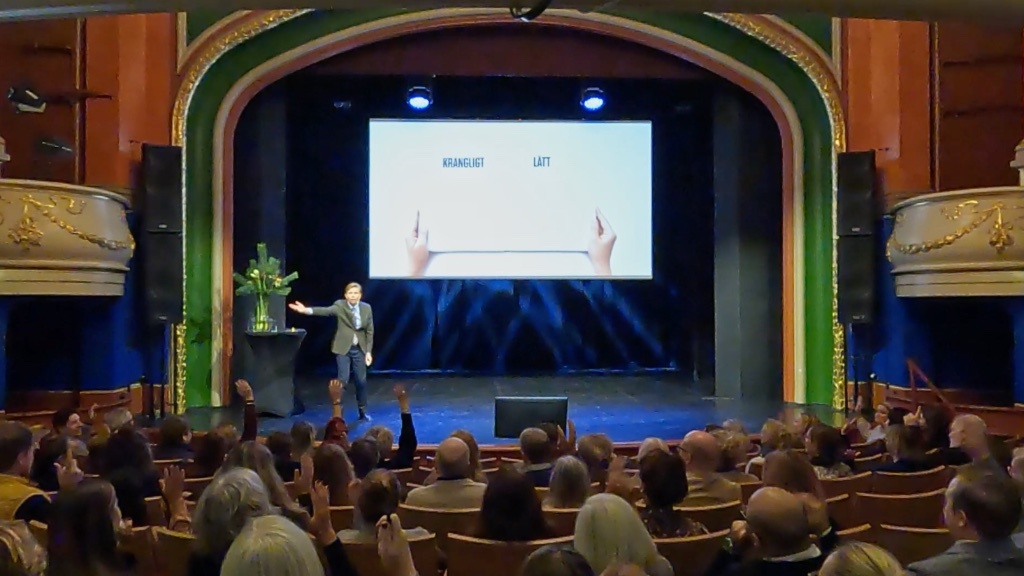 En talare presenterar för en engagerad publik i en teater med orden "KRÅNGLIGT" och "LÄTT" på en skärm bakom honom.