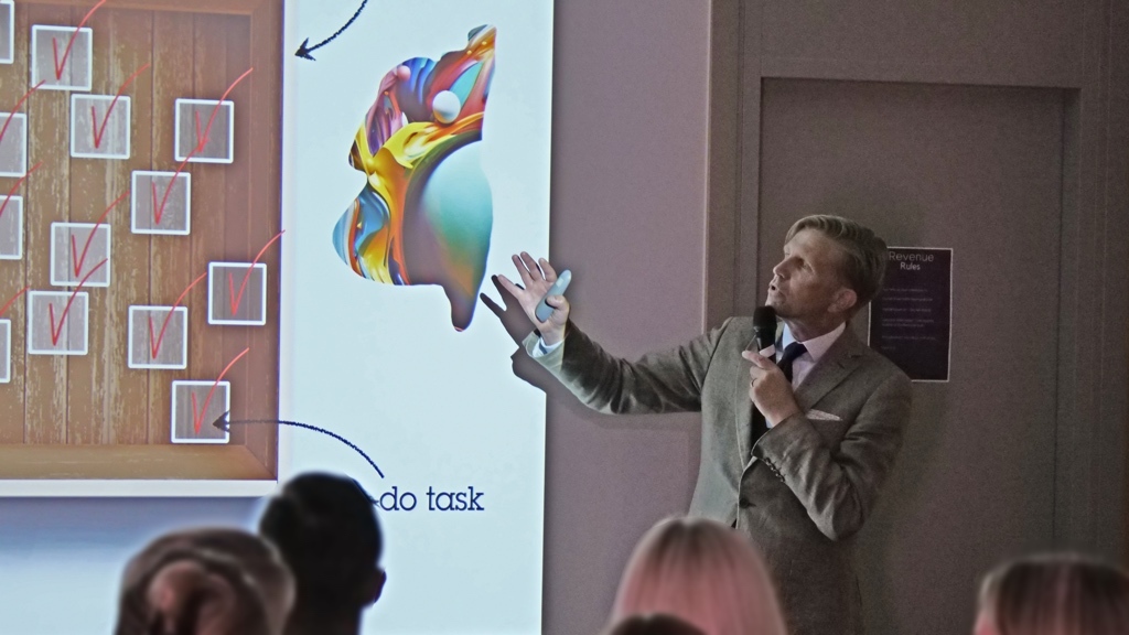 A blonde man in a light brown blazer and blue crocheted tie is lecturing next to a large image with illustrative blobs and to-do tasks in a frame. In the foreground, the audience's heads are visible.
