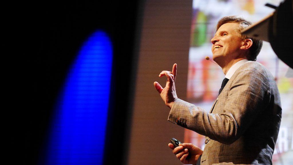 Blonde man in light brown jacket smiles while giving a talk to a large audience, in front of a brightly coloured background.