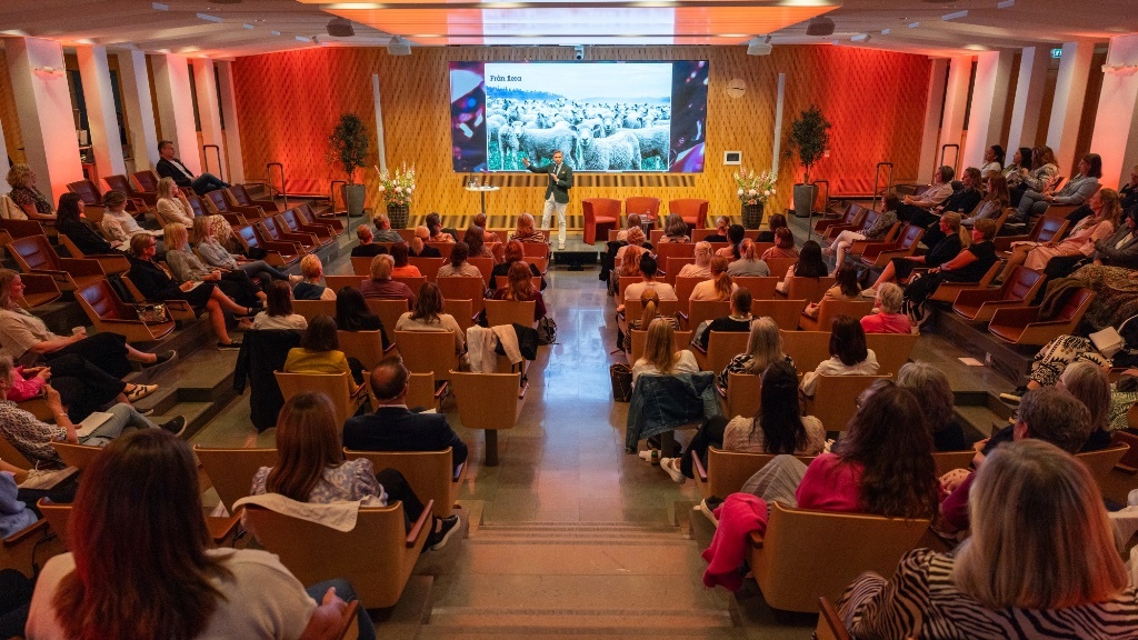 En föreläsare står på scen i en varmt upplyst hörsal och talar inför en stor sittande publik, medan en presentationsbild med får och orden ”Från flera” visas på en stor skärm bakom honom.