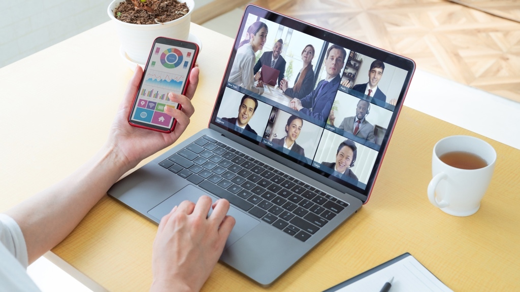 En person deltar i ett videomöte på en laptop samtidigt som de håller en smartphone med färgglada datadiagram, med en kopp te och anteckningsblock på skrivbordet.