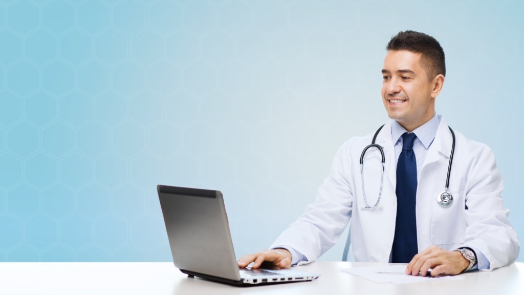 A smiling doctor in a white coat with a stethoscope around his neck is sitting at a desk and working on a laptop.