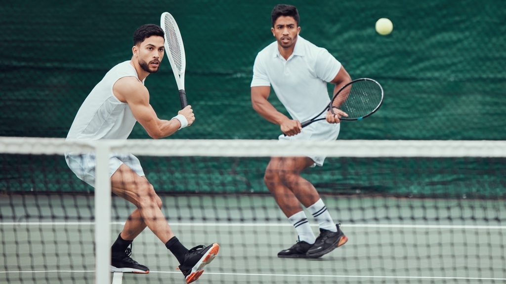 Two men play doubles tennis on a court, both poised to hit the ball with their rackets in an intense match moment.