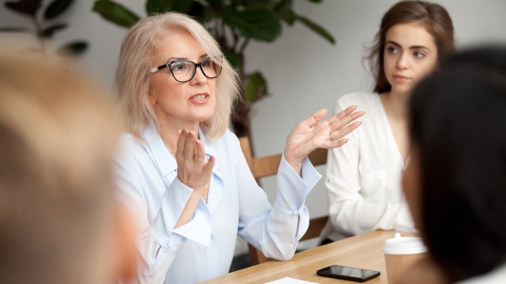 An older woman wearing glasses is speaking passionately during a meeting, with a younger woman and others listening around the table.