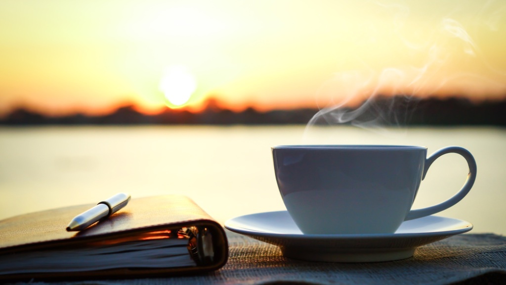 A steaming cup of coffee sits on a wooden surface with a notebook and pen against a sunrise over water.