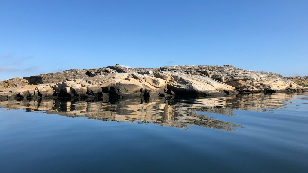 A rocky islet with smooth, sunlit granite is reflected in the calm, mirror-like sea under a clear blue sky.
