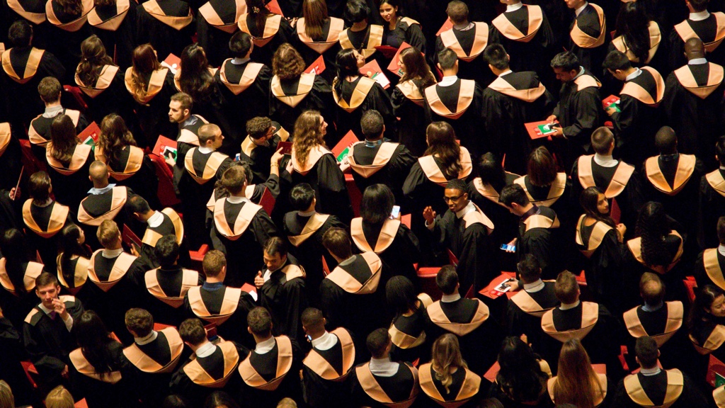 Graduates in ceremonial robes are gathered, some holding flags, during a graduation ceremony.