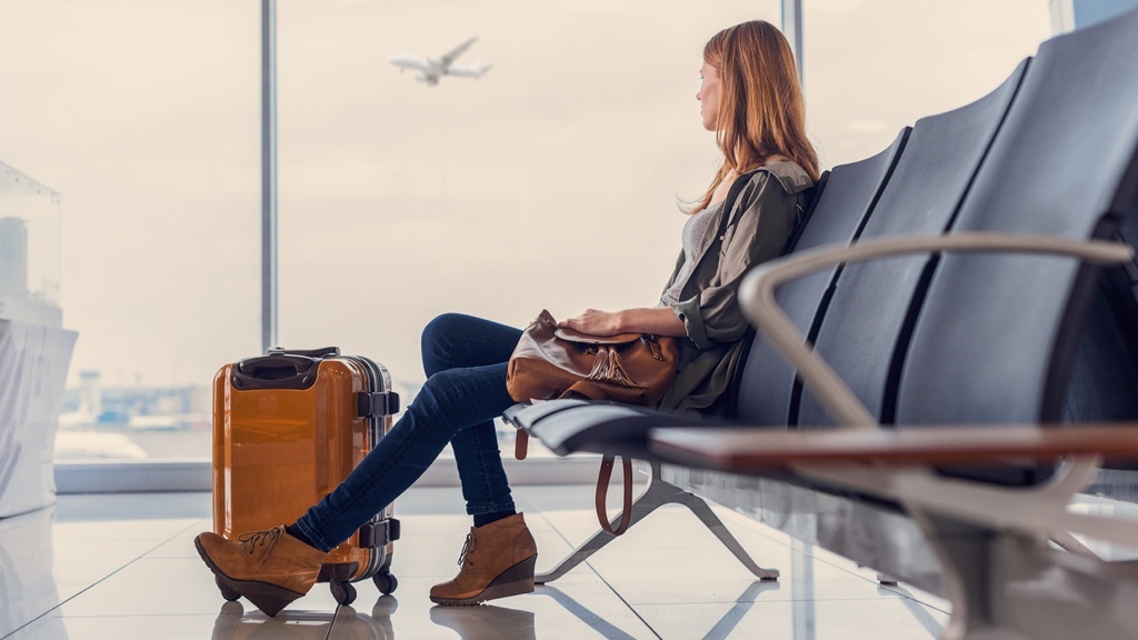 En kvinna sitter i en flygplatsterminal med sitt bagage och tittar på ett flygplan som lyfter utanför fönstret.