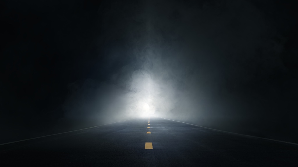 A dark road stretches forward into thick fog, illuminated by a mysterious bright light in the distance.