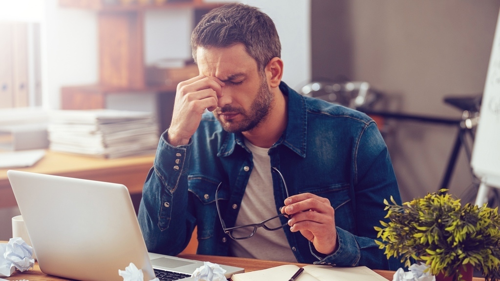 A stressed man sits at a desk with his head in his hand, surrounded by crumpled papers, a laptop, and a notebook.