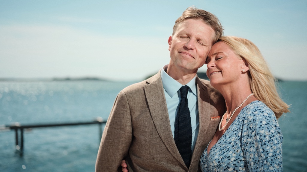 Ett leende par står tätt tillsammans med slutna ögon och njuter av en stilla stund vid havet en solig dag.