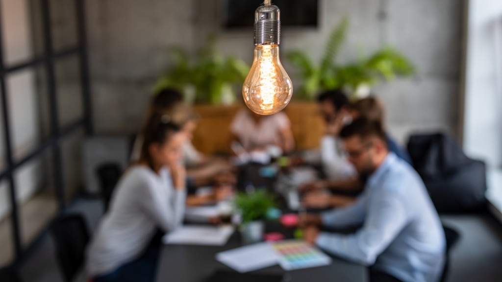 A glowing light bulb hangs in sharp focus above a blurred group of people collaborating around a table, symbolizing a shared moment of creativity or a bright idea during teamwork.