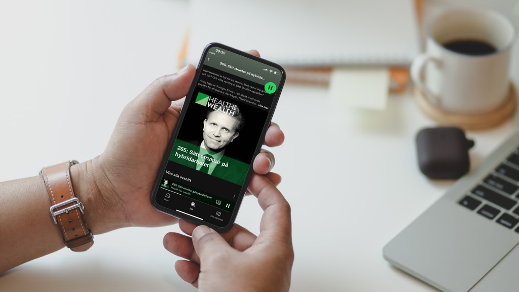 Två händer håller en telefon på vilken ett poddavsnitt spelas i Spotify-appen. I bakgrunden: En kaffekopp och en laptop.