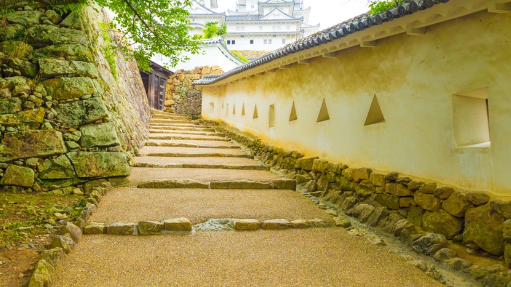 En fridfull stig med stentrappor löper längs med en traditionell vit vägg med triangulära skottgluggar, som leder mot ett slott i bakgrunden.