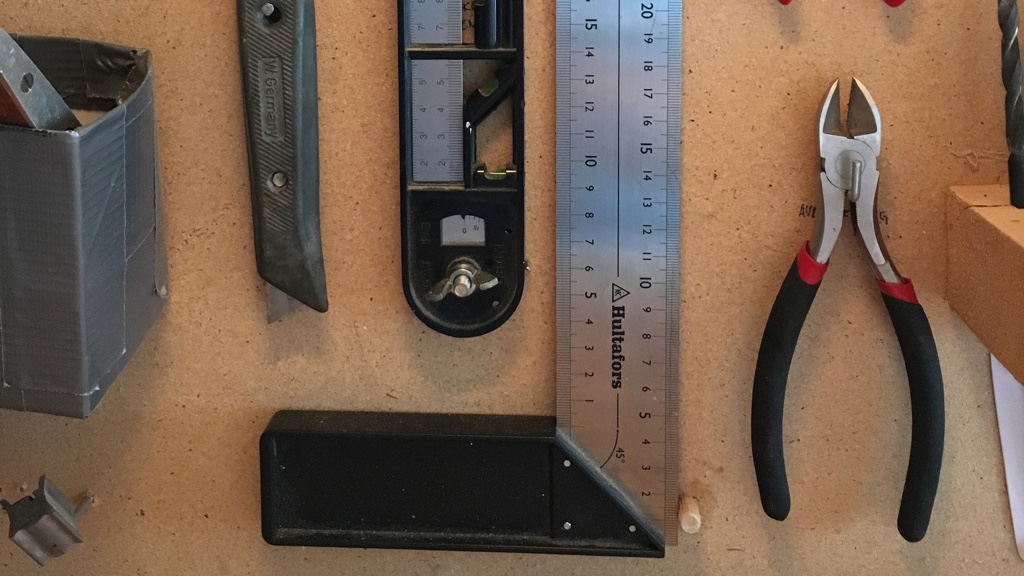 Olika verktyg inklusive en vinkelhake, en tumstock och en tång är organiserade på en verktygstavla.