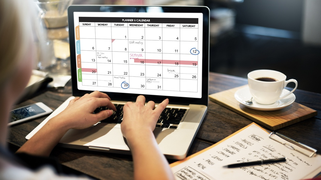 A look over the shoulder of someone typing on her laptop. On the screen is a calendar view. To the right, on the desk sits a coffee cup and a notepad.