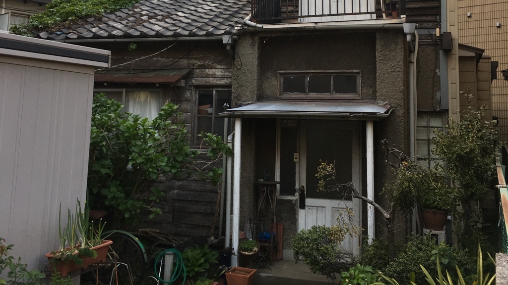 Ett gammalt, slitet japanskt hus med en blandning av trä- och putsade väggar står omgivet av frodig grönska, krukväxter och trädgårdsredskap – vilket ger ett hemtrevligt men något vildvuxet intryck.