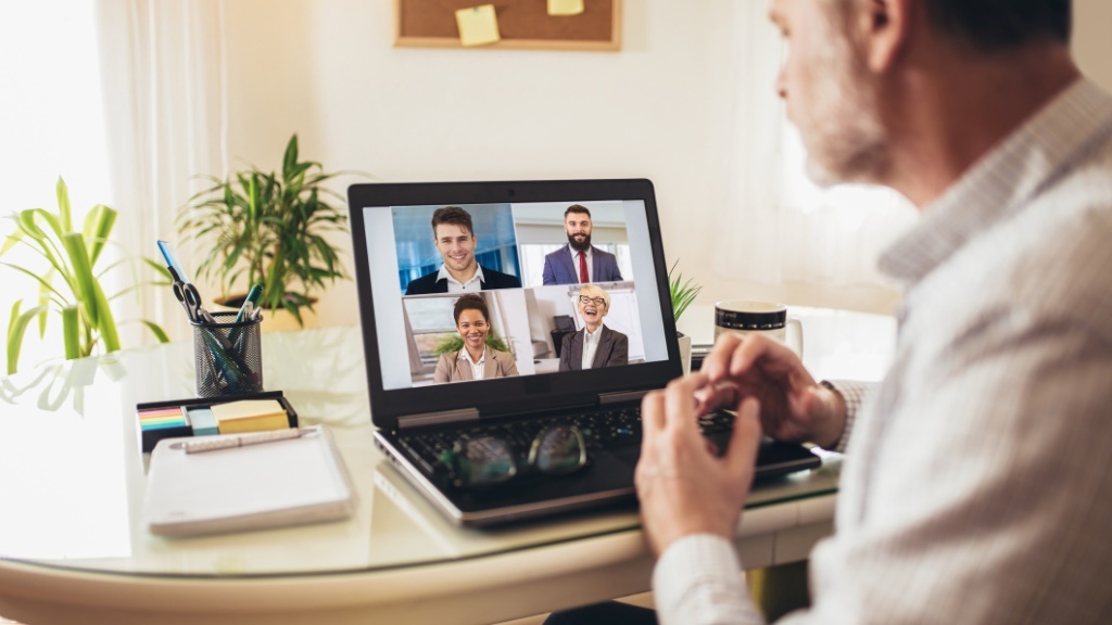 A man sits at a desk in a home office, participating in a video call with four colleagues visible on his laptop screen.
