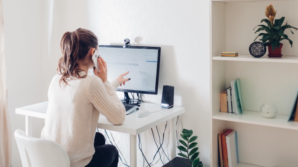 A woman sitting at a desk works on her computer while talking on the phone in a home office.
