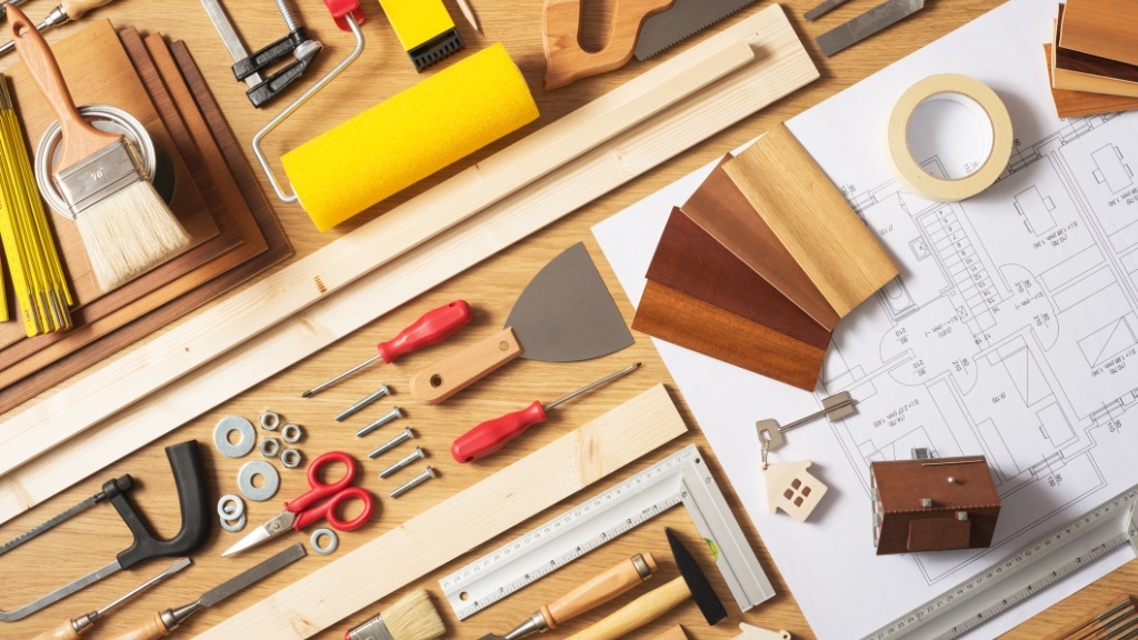 A variety of tools, wood samples, screws, and a house blueprint are neatly arranged on a wooden surface, suggesting a home renovation or interior design project.