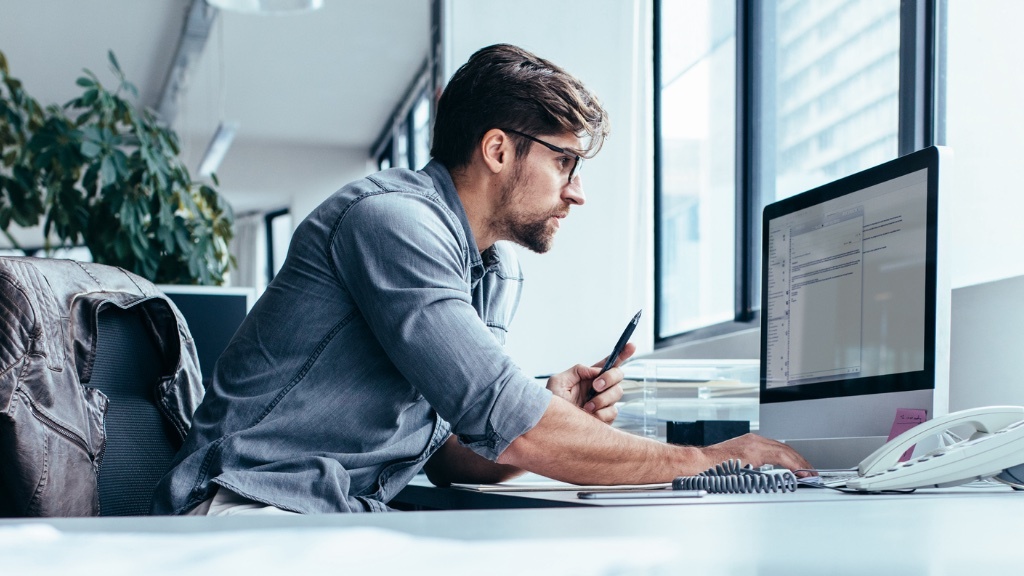 A man wearing glasses and a denim shirt is sitting at a desk, focusing on work on a computer while holding a pen in his hand.
