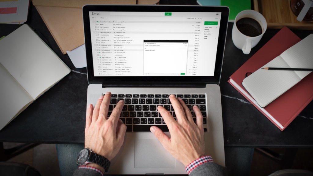 A person is typing on a laptop with an email interface open, surrounded by a notebook, a cup of coffee, and office supplies.