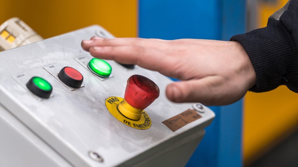 A hand is about to press a red emergency stop button on a control panel with illuminated green and red buttons.