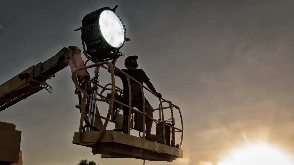 A man is silhouetted against the sky while operating a large spotlight on a crane.