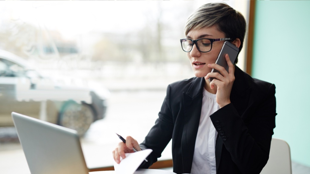 A woman in business attire is talking on the phone while working on a laptop and taking notes at a desk.