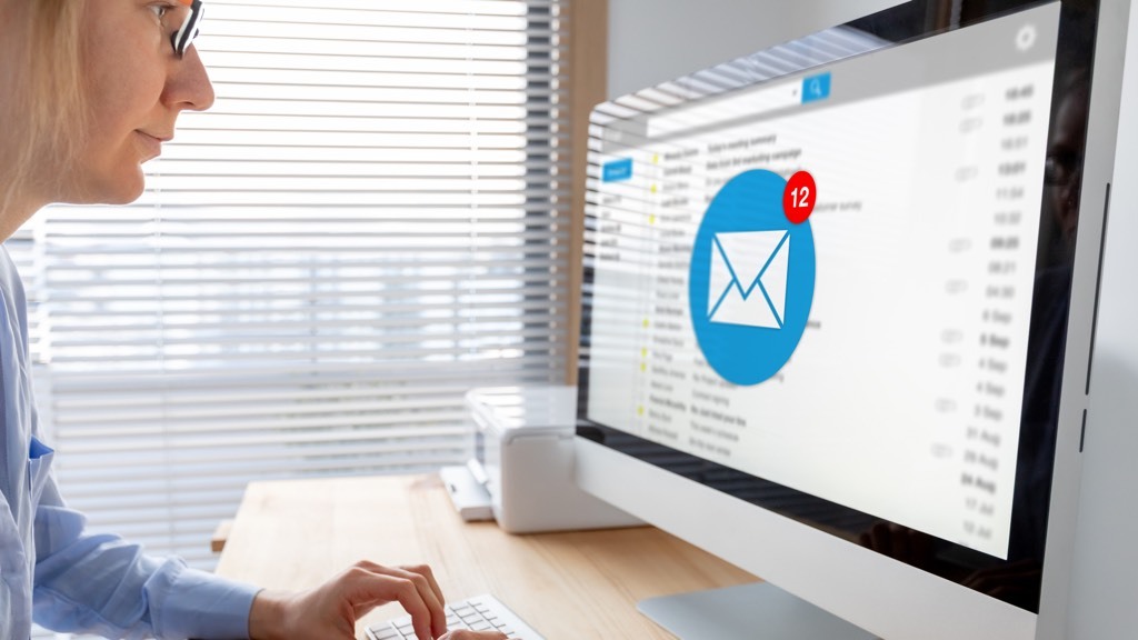 En kvinna sitter vid ett skrivbord och kollar sin e-post på en stationär dator, med en avisering som visar 12 nya meddelanden.