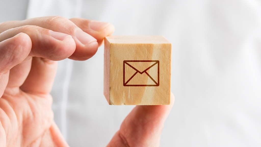 A hand holds a wood block cube with an envelope stamp on one side, in front of a white shirt chest.