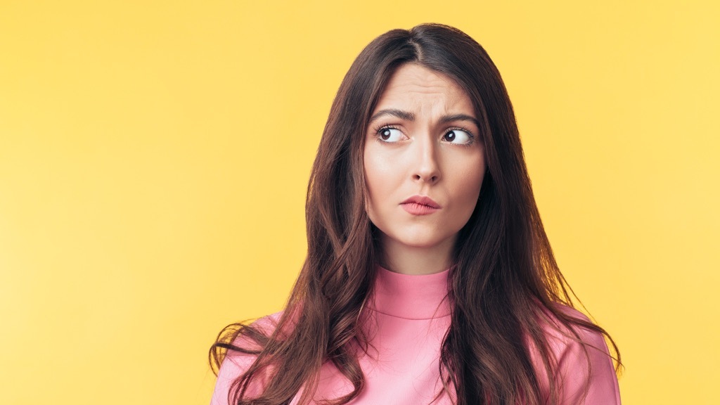 A young woman with long brown hair wearing a pink top looks to the side with a puzzled or skeptical expression against a yellow background.