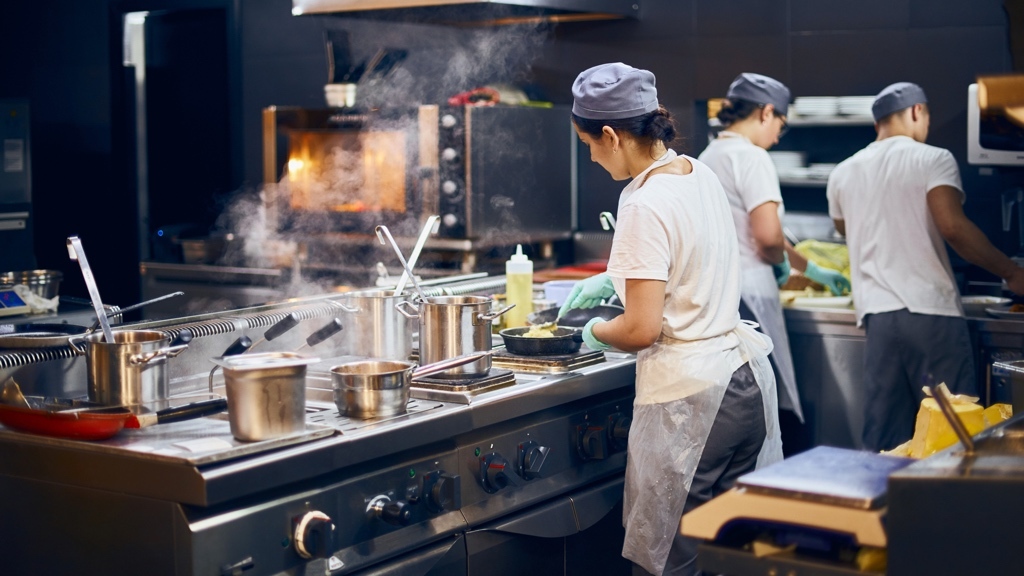 Tre restaurangkockar arbetar i ett modernt restaurangkök. Det står kastruller på spisen.