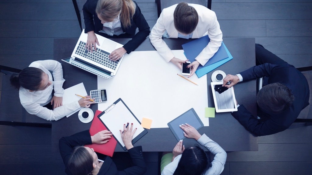 A group of professionals is gathered around a table in a meeting, working on various tasks with laptops, tablets, notepads, and a calculator.