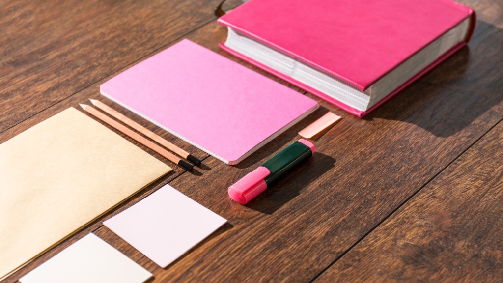Ett urval av kontorsmaterial inklusive en rosa anteckningsbok, två pennor, en rosa post-it-block, en tuschpenna och vanliga kort utlagda på en träyta.