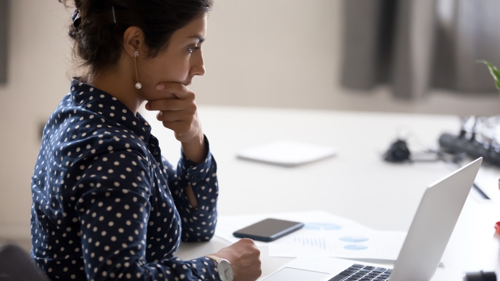A woman in a navy polka-dot shirt is sitting at a desk, focused and thoughtful, as she looks at her laptop screen with her hand resting on her chin.