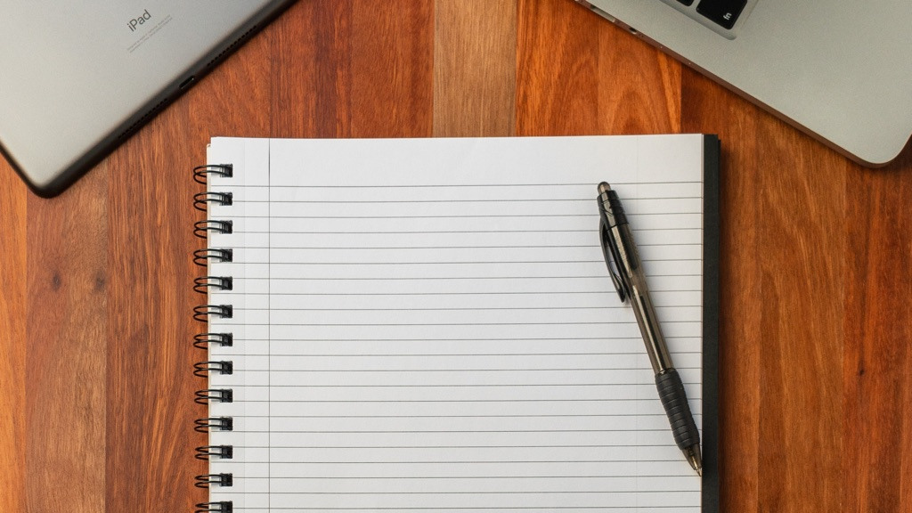 Notepad with an in rollerball pen, placed on a wooden desk. An iPad in the top left corner and a glimpse of a MacBook in the top right.