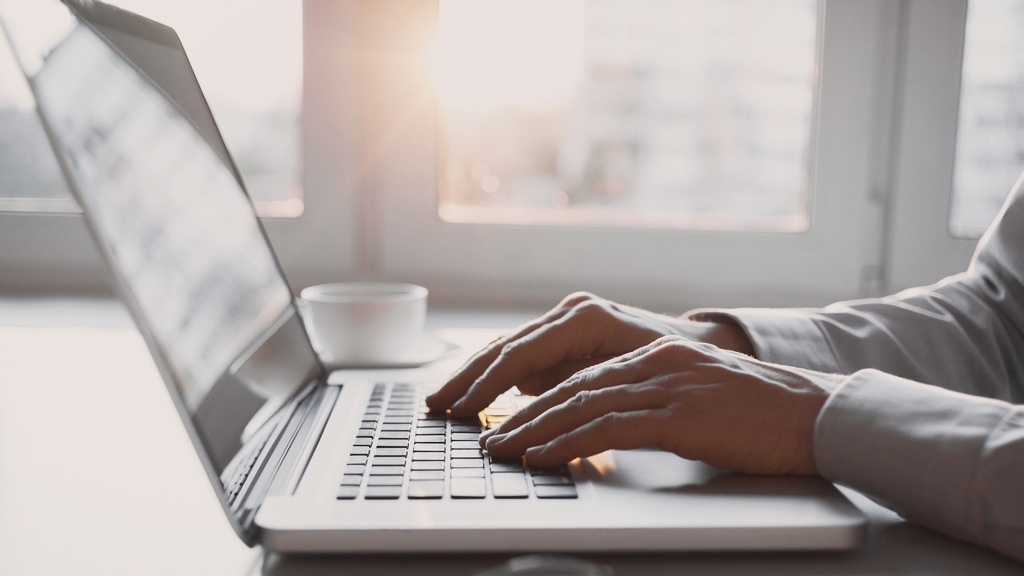 En person skriver på en bärbar dator i ett ljust rum med solljus som strömmar in genom fönstret, och en kaffekopp står bredvid.