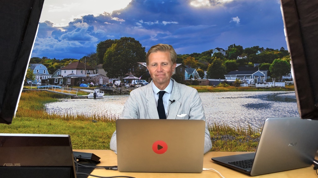 A blonde man in a light grey seersucker jacket and a blue knitted tie sits at his laptop in front of a beautiful coastal scene from Massachusetts.