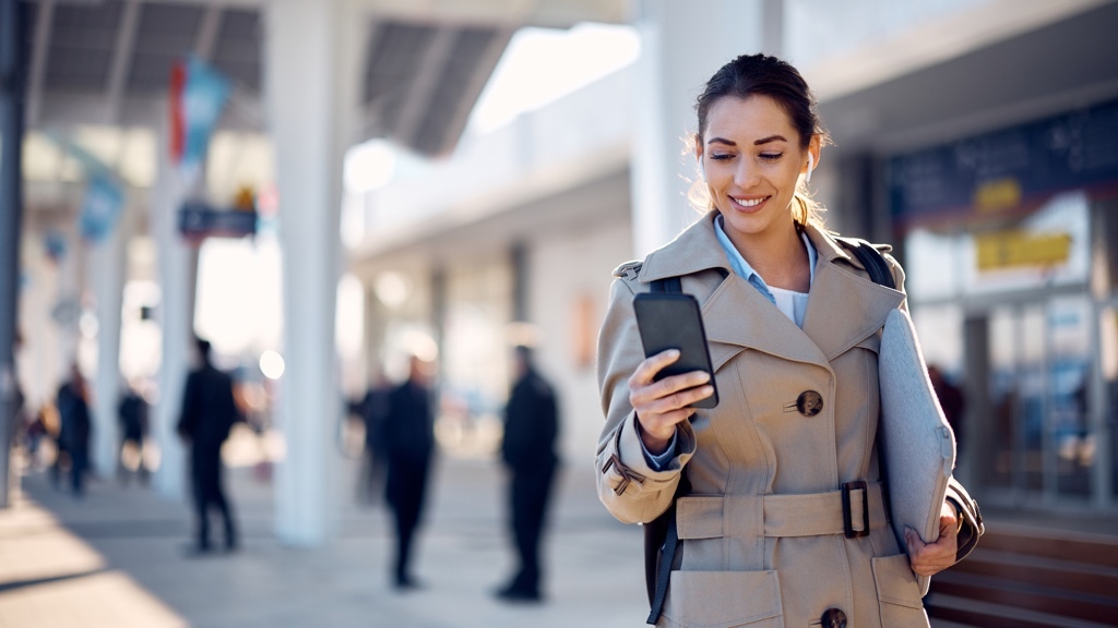 A smiling woman in a trench coat looks at her smartphone while standing outdoors with a laptop under her arm and blurred figures in the background.